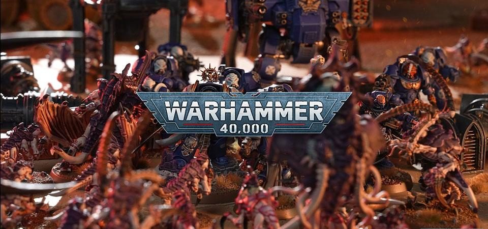 El logo de Warhammer 4000 superpuesto a una imagen de batalla de miniaturas que enfrenta a exterminadores y un dreadnought ballistus contra una fuerza (en primer plano y de espaldas) de tiránidos formada por termagantes, saltadores de Von Ryan y un neurotirano.